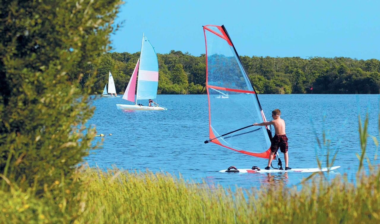 Jonge windsurfer en zeilboten op het meer bij camping LIBRANOO Naturiste Arnaoutchot, Landes, Frankrijk.