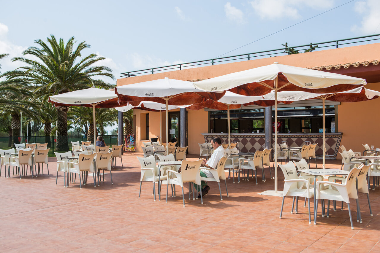 Buiten terras van restaurant-bar met tafels, parasols en palmbomen op camping CAPFUN Aube in Deltebre, Tarragona (43).