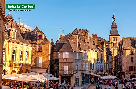 Levendig plein in Sarlat-la-Canéda met historische gebouwen en restaurantterrassen.
