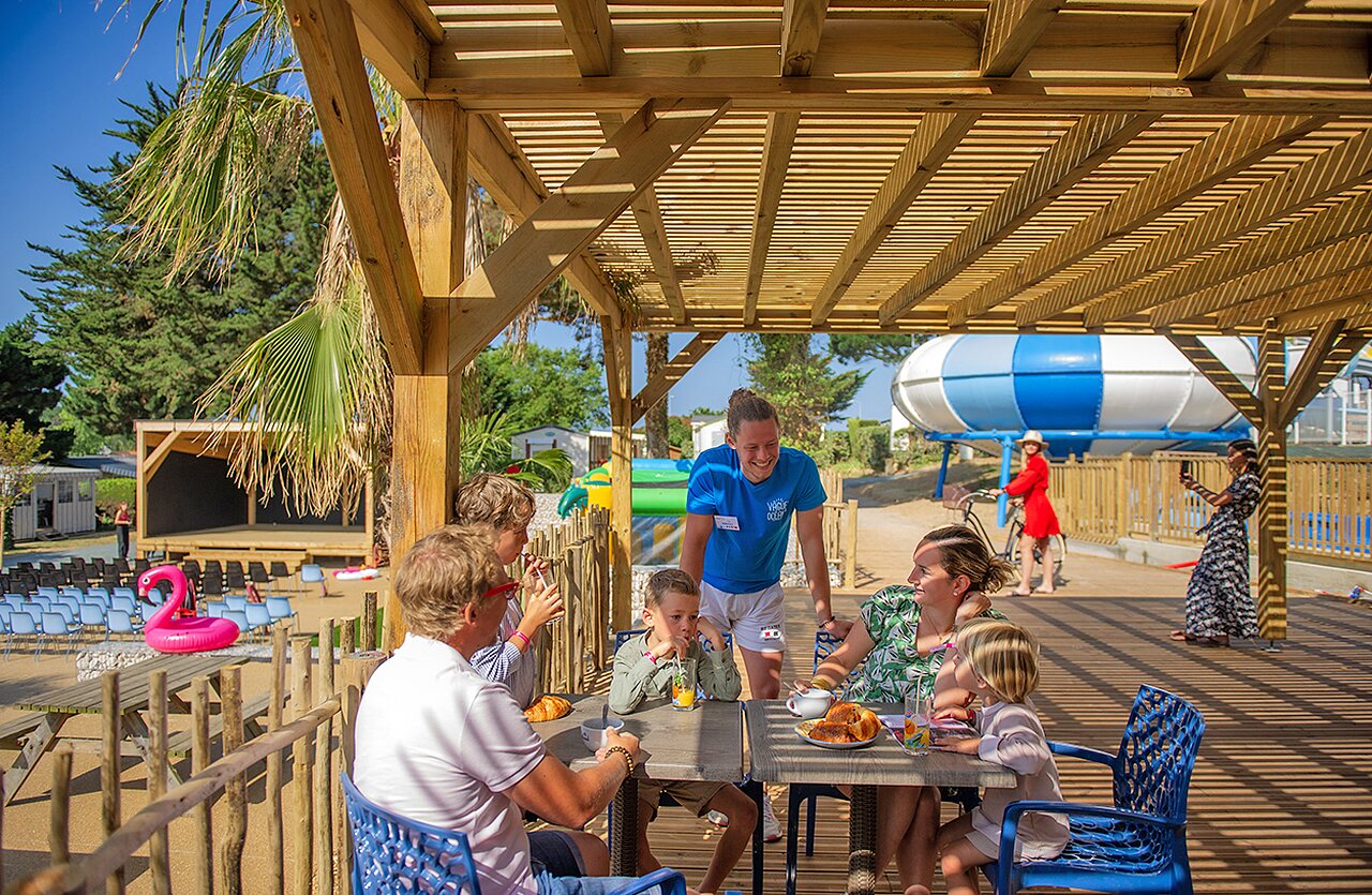 Familie ontbijtend onder pergola, animator, podium, glijbaan op camping CLICOCHIC Brandais in Brem sur Mer (85).