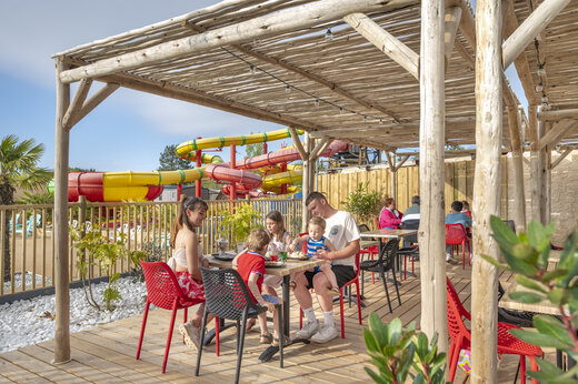 Familie luncht op restaurantterras, waterglijbanen zichtbaar op camping CAPFUN Chateau d'Arvid (14).