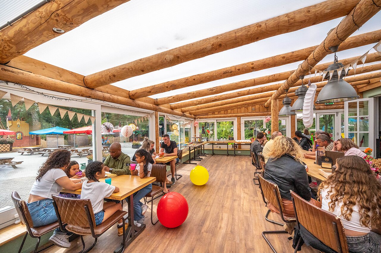 Families en kinderen dineren in overdekt restaurant met uitzicht op speeltuin op camping CAPFUN De Scheepsbel in Doornspijk.