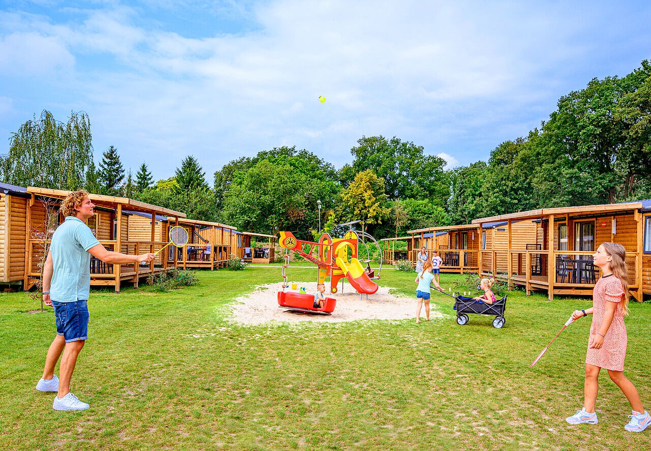 Familie speelt badminton en kinderen in speeltuin, houten accommodaties op camping CAPFUN Eibernest in Eibergen.