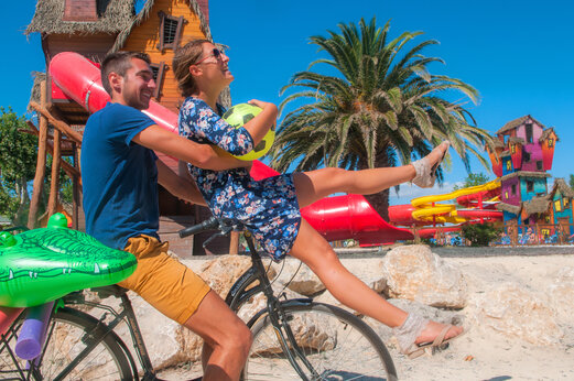 Vrolijk stel op fiets met zwembanden, kleurrijke waterglijbanen op camping CAPFUN Féerix in MARSEILLAN PLAGE.