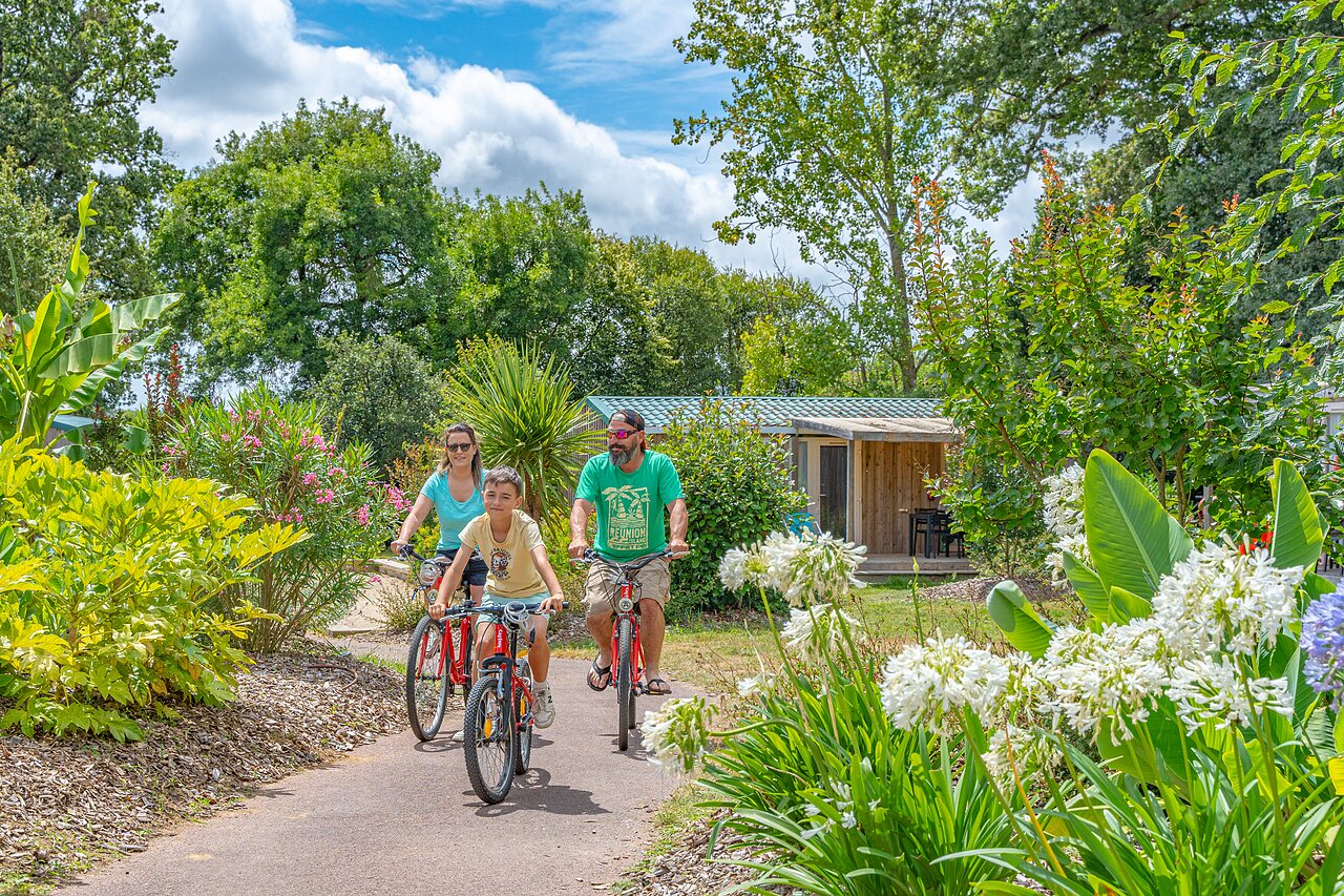 Familie fietst op bloemrijk pad, accommodatie op achtergrond op camping CAPFUN Forges in Avrille (85).