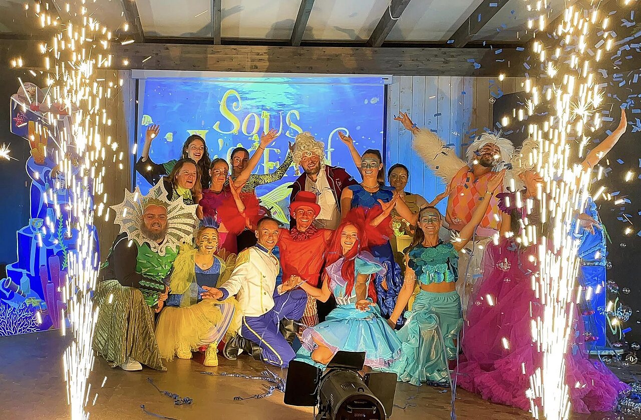 Artiesten in kleurrijke kostuums op podium met pyrotechnische effecten op camping CLICOCHIC Longchamp in Saint Lunaire (35).