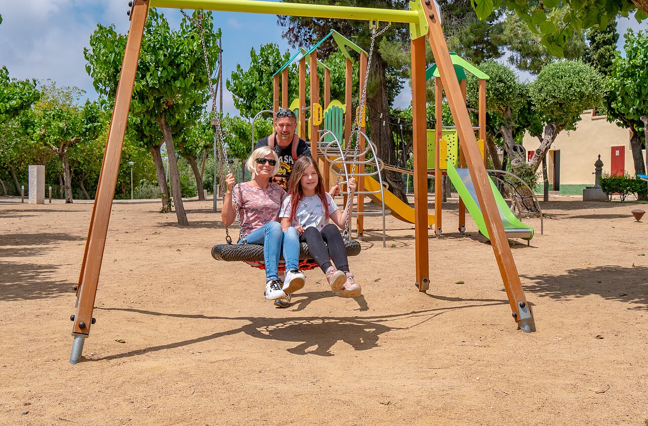 Familie op de schommel van de speeltuin op camping CAPFUN Mirmanda in Cambrils.